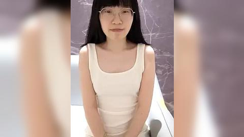 Media: Video of an Asian woman with long black hair and fair skin, wearing glasses and a white tank top, sitting against a marble wall.