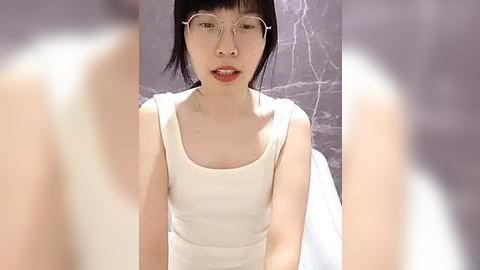 Media: Video of an Asian woman with short black hair, wearing glasses, and a white tank top, sitting in a room with gray marble walls.