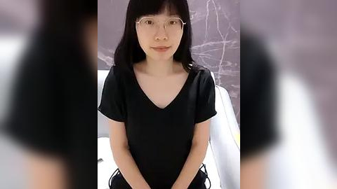 Media: Video of a young East Asian woman with straight black hair, wearing clear glasses and a black V-neck shirt, sitting against a textured gray wall, blurred background.