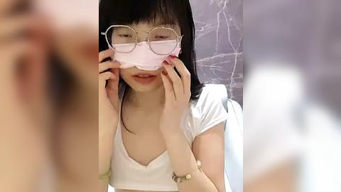 Media: Video of an Asian woman with straight black hair, wearing glasses and a pink surgical mask, sitting in a white chair. She has fair skin and is wearing a white top. Background shows blurred marble pattern.