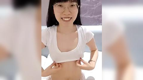 Media: A video of an Asian woman with long black hair, glasses, and a white crop top, standing in a modern room with a gray brick wall and white bedding.