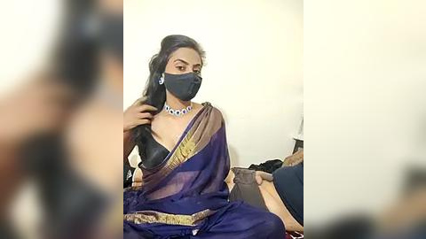 Media: A video shows a woman with medium skin tone and long, dark hair, wearing a black face mask, black blouse, and a blue sari with gold trim. She sits against a plain white wall, with blurred figures in the background.