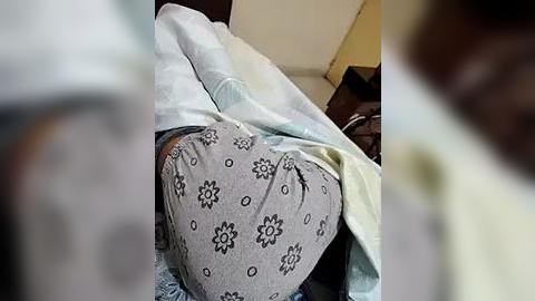 Media: Video of a person lying on a bed, wearing light gray pajamas with black flower patterns, partially covered by a white sheet and a light blue blanket. The room has beige walls and a brown chair in the background.