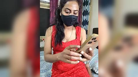 Media: Video of a South Asian woman with medium-brown skin, wearing a red lace dress, black face mask, and long black hair, holding a phone in a bedroom with patterned curtains and a blue bedspread.