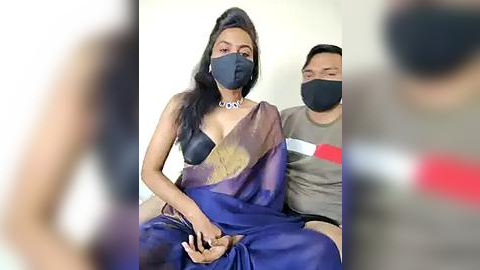 Media: Video of a woman wearing a blue sari with a black mask, seated between two men, one in a grey shirt, in a plain indoor setting.