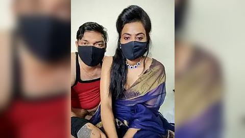 Media: Video of two South Asian individuals wearing black face masks, sitting closely, one in a red tank top, the other in a blue sari, with blurred background.