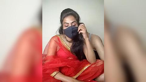 Media: Video of a South Asian woman with medium skin tone, wearing a red saree with gold trim, black mask, and dark hair, sitting with eyes closed, holding a smartphone, against a plain light-colored background.