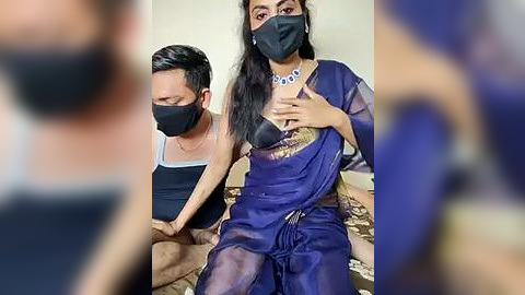 Media: A video shows a man in a black tank top and face mask, sitting next to a woman in a blue sari, black face mask, and black necklace, with a blurred figure in the foreground.