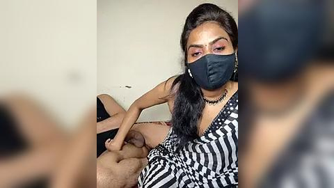 Media: Video: A woman with dark skin, wearing a black mask and striped dress, is seen in a close-up, holding a man's erect penis. Background is plain, with blurred figures.