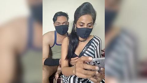 Media: A video captures a man in a blue tank top and black mask sitting behind a woman in a striped dress, both wearing black masks, focusing on a smartphone.