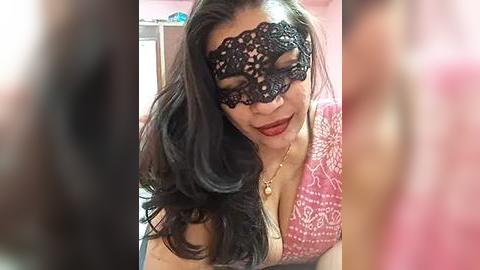 Media: Video of a woman with long black hair, wearing a black lace masquerade mask, red lipstick, and a pink patterned top, smiling. Background blurred, indoor setting.