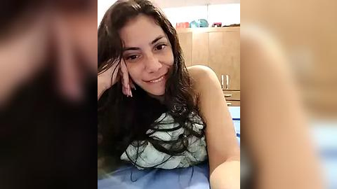 Media: Video of a smiling woman with long dark hair, wearing a black strapless top, lying on a bed, blurred background with wooden furniture.