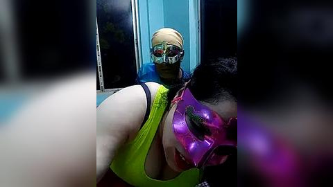 Media: Video of two women in vibrant, colorful masks: one in a green tank top, the other in a purple mask, both looking at a blonde woman in a blue mask and goggles.