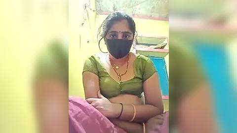 Media: A video of a woman with medium brown skin, wearing a green blouse, black mask, and gold jewelry, sitting on a bed in a yellow room.