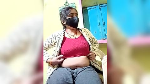 Media: Video of a pregnant woman in a bright room, wearing a black mask, red crop top, and blue pajama pants, sitting with her hands on her belly, next to a window with blue shutters.