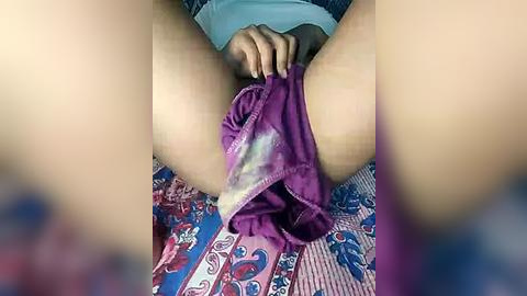 Media: A video showing a person in a purple shirt with a light-colored undershirt, sitting on a patterned bedspread, pulling up purple shorts that are stained with white liquid, revealing bare skin.