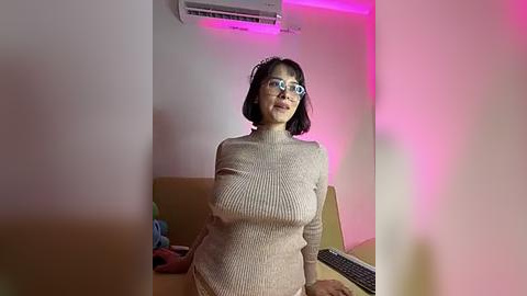 Media: A video of an Asian woman with short black hair, wearing glasses and a beige ribbed sweater, standing indoors with a TV and couch in the background, illuminated by pink ambient lighting.