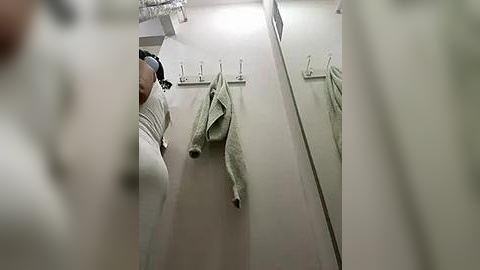 Media: Video of a person in a white long-sleeve shirt, partially obscured by a mirror, hanging towels on a wall in a bathroom with a beige color scheme.