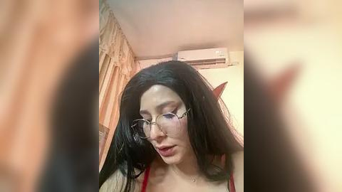 Media: Video of a fair-skinned, slender woman with long black hair, wearing glasses, a red top, and a worried expression. She stands in a beige room with beige curtains and an air conditioner on the wall.