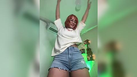 Media: Video of a red-haired woman with fair skin, wearing a white t-shirt and high-waisted denim shorts, arms raised, in a dimly lit room with green light and decorative bottles.