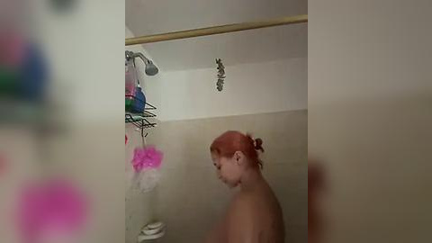 Media: Video of a young, fair-skinned girl with red hair in a shower. She is topless, looking downward. The shower has a metallic mesh shelf with pink and purple soaps, and a beige tiled wall.