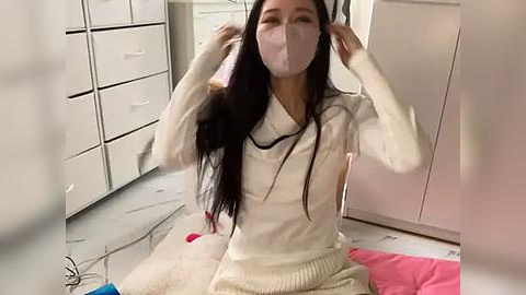 Media: Video of an Asian woman with long black hair, wearing a white sweater and face mask, adjusting headphones while seated on a pink blanket in a tidy, modern bedroom with white cabinets and a beige carpet.