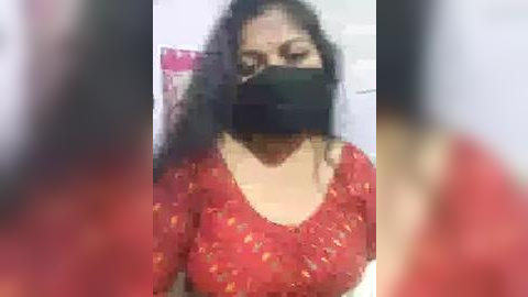 Media: Video of a woman with long black hair, wearing a red floral-patterned blouse, black face mask, and black face covering, standing indoors with blurred background.