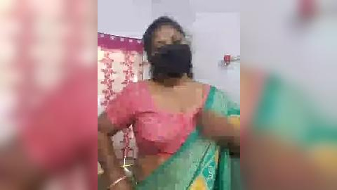 Media: A blurred video shows a woman in a pink blouse and green sari with a black mask, standing in a room with a red curtain and white walls.