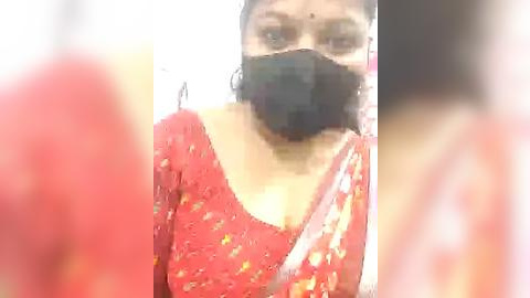 Media: Video of a woman wearing a red floral-patterned saree, black face mask, and black hair, standing indoors with blurred background.