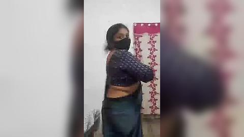 Media: A blurry video of a woman wearing a black mask, blue jeans, and a textured black sweater, standing in a dimly lit room with a floral curtain and white walls.