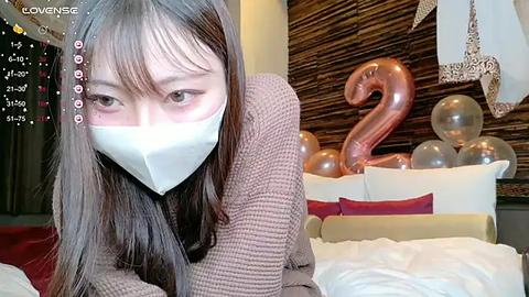 Media: A video of an Asian woman with long black hair, wearing a white face mask, brown sweater, and brown pants, lying on a bed with white sheets and a \"2\" made of balloons in the background.