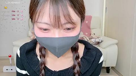 Media: Video of an Asian woman with light skin and brown hair in braids, wearing a gray duct tape gag and a black sweater, kneeling in a modern living room with a white couch and pink pillow.