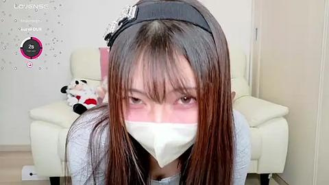 Media: A video of an Asian woman with long brown hair, wearing a black headband and a white face mask, kneeling in a living room with a white chair and a plush toy.