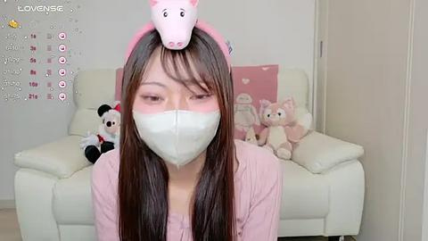 Media: Video of an East Asian woman with long brown hair and a pink pig headband, wearing a white face mask, in a pink outfit, sitting on a white couch with stuffed toys, calendar on wall.