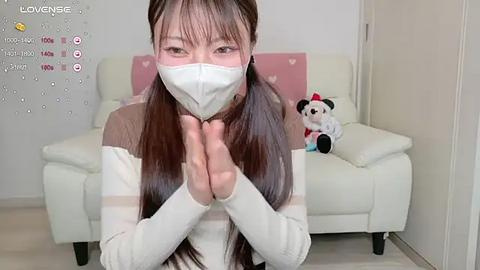 Media: A video of an Asian woman in a white mask, white sweater, and black pants, kneeling and praying in a modern, minimalist living room with a white couch and a stuffed panda.