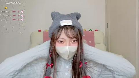 Media: A video of an Asian woman with long brown hair, wearing a gray cat hat and a white mask, lying on a bed with light blue bedding and a pink pillow.