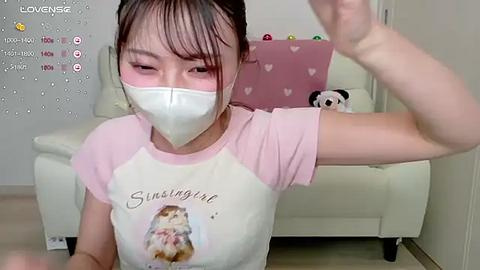 Media: Video of an Asian woman with light skin, wearing a white face mask, pink and white T-shirt, and holding a yoga pose in a living room.