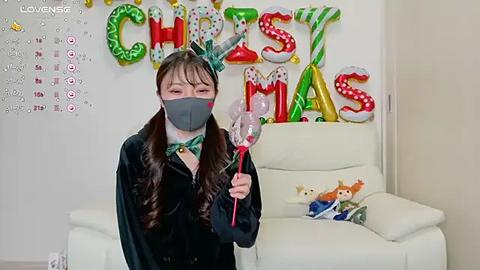 Media: Video of an Asian woman in a black bunny costume, holding a red wand, with a face mask and green bow tie. Christmas decorations and a stuffed animal on a white couch in the background.