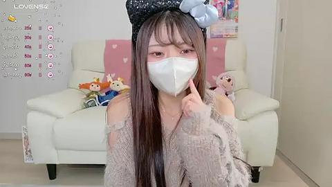 Media: Video of an Asian woman in a beige sweater, wearing a black hat and white mask, sitting on a white couch with plush toys in a light-colored room.