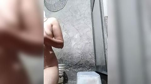 Media: Video of a nude person with fair skin and medium build, standing in a small, dimly lit bathroom with a metallic showerhead, a toilet, and a plastic bin on the floor.