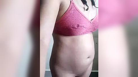 Media: A video of a pregnant woman with light skin, wearing a pink sports bra, standing against a blurred background. Her belly is prominent, and her dark hair is visible at the top of the image.