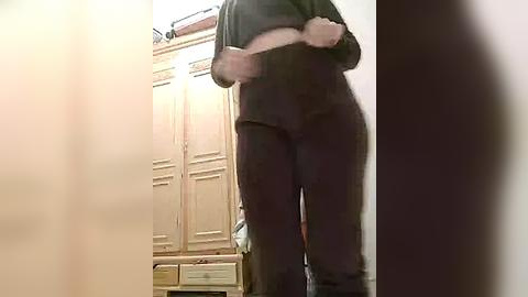 Media: A video captures a person from the waist down, wearing dark pants, standing in front of a wooden cabinet with drawers, in a dimly lit room.