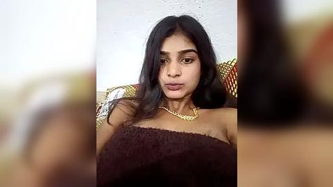 Media: Video of a young South Asian woman with long black hair, wearing a brown knitted sweater, sitting on a patterned couch against a white wall, with blurred background.