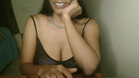 Media: Video of a smiling woman with medium brown skin, long black hair, wearing a black sequined halter top, holding her chin, in a dimly lit room.