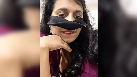 Media: Video of a South Asian woman with long black hair, wearing a black face mask, lying on a bed. Her face is partially covered, and she has a faint smile.