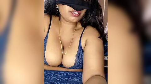 Media: A close-up video of a woman with medium brown skin and black hair, wearing a blue lace bra, black blindfold, and a gold necklace, in a dimly lit room.