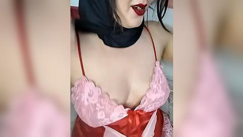 Media: Video of a light-skinned woman with dark hair, wearing a red satin bra with pink lace trim, black choker, and red lipstick, sitting on a red couch.