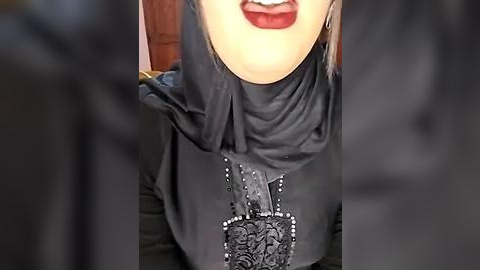 Media: A close-up video of a person with light skin wearing a black hijab adorned with intricate silver embroidery and a matching necklace, set against a blurred background.