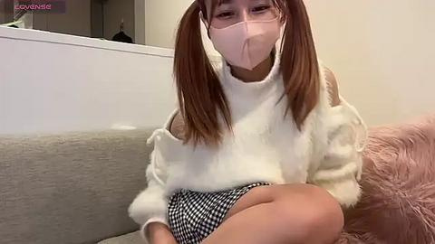 Media: Video of an East Asian woman with light skin and pigtails, wearing a white turtleneck sweater and black and white checkered skirt, sitting on a grey couch in a modern living room.