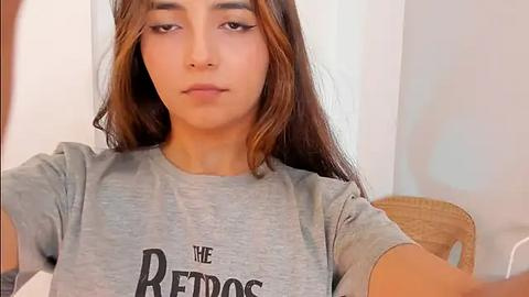 Media: Video of a young woman with medium skin tone, long brown hair, wearing a grey T-shirt with \"THE RED DOG\" in bold letters. She has a neutral expression, sitting indoors against a plain white wall.